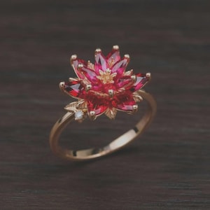 Puede incluir: Un anillo de oro con un diseño de flor de varias capas compuesto por piedras preciosas de color rojo intenso con talla marquesa. El centro de la flor tiene estambres dorados y una pequeña piedra preciosa redonda y clara en el aro.