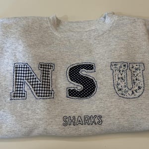 Puede incluir: Una sudadera de cuello redondo en color gris jaspeado claro con las letras NSU aplicadas en diferentes telas: una N de cuadros vichy en blanco y negro, una S de lunares en blanco y negro y una U floral en azul y blanco. La palabra SHARKS está bordada debajo.