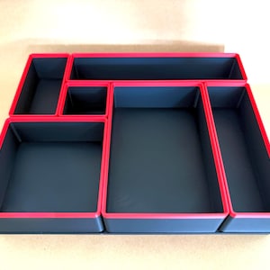 May include: A rectangular drawer organizer tray featuring a dark gray interior and vibrant red edges. The tray is divided into six distinct compartments of varying sizes, providing structured storage space for organizing small items.