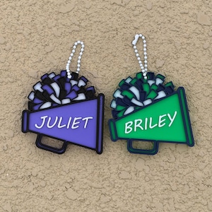 May include: Two cheerleading megaphone ornaments with attached white beaded chains. One megaphone is purple with the name JULIET in white and a black, white, and purple pom-pom. The other is green with the name BRILEY in white and a black, white, and green pom-pom.