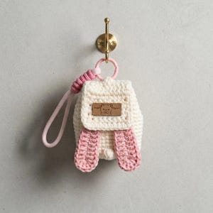 以下が含まれることがあります： クリーム色のクロシェ編みミニリュック型キーホルダー。ピンクのアクセントとして、2つの長いピンクの耳とピンクのリストストラップが付いています。フロントフラップには、クマのイラストと「Bear Clever」という文字が刻まれた茶色のレザー調の長方形パッチがあしらわれています。