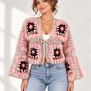 May include: A cropped crochet cardigan featuring a patchwork of square motifs in shades of pink, cream, and dark maroon. The garment includes long, flared sleeves, a textured fuzzy yarn border, and a thin crocheted tie closure at the centre front.