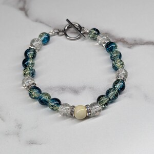 May include: A beaded bracelet featuring a mix of translucent blue and clear crackle glass beads, accented with small silver-toned spacers and a single smooth, cream-coloured central bead. The bracelet is secured with a silver-toned metal toggle clasp.