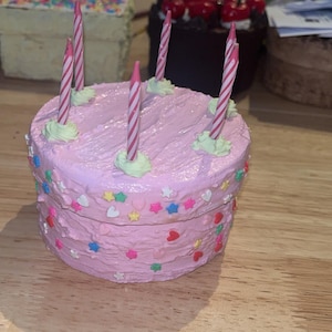 May include: A round pink cake-shaped trinket box with textured frosting, decorated with colorful star and heart-shaped sprinkles. Five thin pink and white striped birthday candles are inserted into the top, each resting on a small dollop of light green icing.