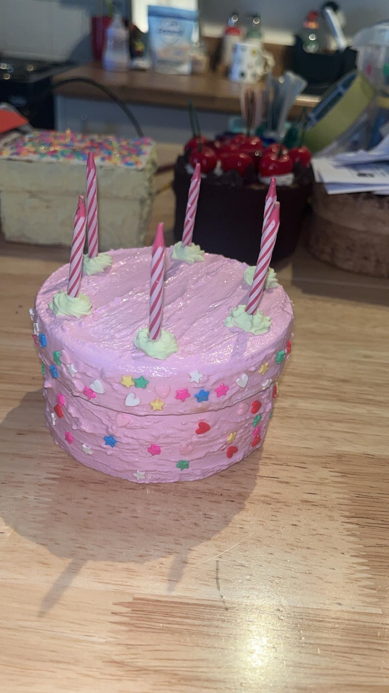 Pu&ograve; includere: Un portagioie rotondo a forma di torta con glassa testurizzata, decorato con zuccherini colorati a forma di stelle e cuori. Cinque sottili candeline di compleanno a righe rosa e bianche sono inserite sulla parte superiore, ognuna su un piccolo ciuffo di glassa verde chiaro.