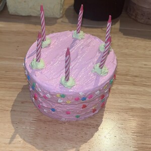 Pu&ograve; includere: Una scatola rotonda a forma di torta con glassa rosa, dotata di cinque candeline di compleanno a righe sulla parte superiore. I lati della scatola sono decorati con piccoli zuccherini colorati a forma di stella e cuore incastonati nella glassa.