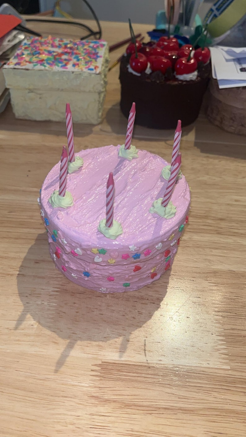 Pu&ograve; includere: Una scatola rotonda a forma di torta con glassa rosa, dotata di cinque candeline di compleanno a righe sulla parte superiore. I lati della scatola sono decorati con piccoli zuccherini colorati a forma di stella e cuore incastonati nella glassa.