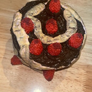K&ouml;nnte beinhalten: Eine runde Schokoladenkuchen-Skulptur mit dunkelbraunem Zuckerguss, einem geschwungenen beigen Dekorelement und mehreren texturierten, roten, himbeerartigen Verzierungen, die auf der Oberfl&auml;che und an den Seiten des Kuchens angeordnet sind.