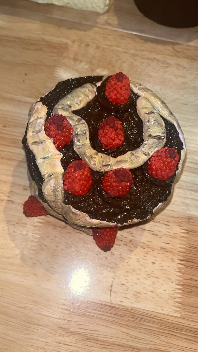 K&ouml;nnte beinhalten: Eine runde Schokoladenkuchen-Skulptur mit dunkelbraunem Zuckerguss, einem geschwungenen beigen Dekorelement und mehreren texturierten, roten, himbeerartigen Verzierungen, die auf der Oberfl&auml;che und an den Seiten des Kuchens angeordnet sind.