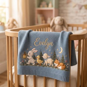 May include: A dusty blue knit baby blanket featuring the embroidered name Evelyn in gold script. The design includes a rabbit holding a lantern, a yellow duckling, a white lamb, and a fox among pine trees, flowers, and a crescent moon.
