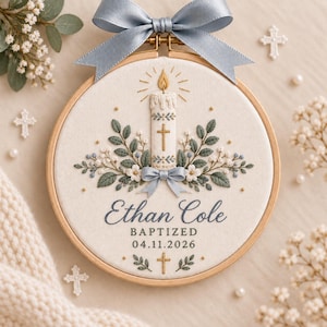 Puede incluir: Un bastidor de bordado circular de madera con tela color crema que presenta un diseño bordado a mano de una vela encendida, hojas verdes y flores blancas. Un lazo de cinta azul claro está sujeto en la parte superior. El texto dice Ethan Cole BAPTIZED 04.11.2026.