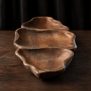 Puede incluir: Una bandeja de servicio de madera de tres secciones tallada en madera de nogal marrón oscuro con una forma orgánica natural e irregular. La bandeja cuenta con tres compartimentos distintos en forma de cuenco con vetas de madera visibles.