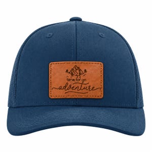 May include: A navy blue trucker hat featuring a rectangular tan faux leather patch on the front. The patch displays a black graphic of mountains and trees above the text "time for an adventure" written in a cursive script font.