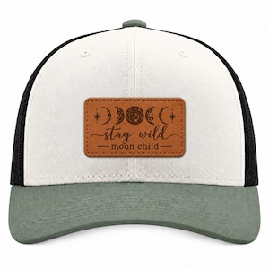 May include: A white and black trucker hat with a sage green brim. Centered on the front is a rectangular brown patch featuring an engraved moon phase design, stars, and the text "stay wild moon child" in a cursive and sans-serif font.