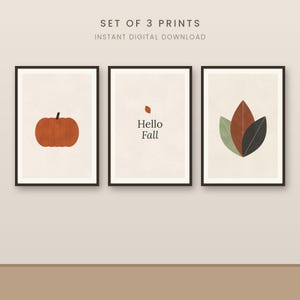 May include: A set of three digital wall art prints featuring a pumpkin, the text Hello Fall with a small leaf, and a cluster of three leaves in green, brown, and dark grey. Each print is framed in black and set against a light beige background.