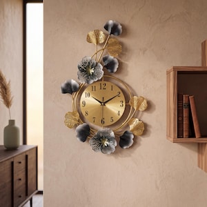 May include: A wall clock featuring a circular gold face with black numerals and hands, surrounded by a metal frame decorated with gold and dark gray ginkgo leaf and flower motifs arranged in an asymmetrical vertical pattern.