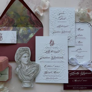 May include: A wedding stationery set featuring a deep red envelope with a floral liner, white embossed cards with black calligraphy, a die-cut bust illustration, a small pink velvet ring box with a diamond ring, and a white ribbon.