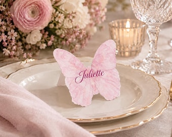 Può includere: Un segnaposto a forma di farfalla in carta rosa chiaro con una texture ad acquerello. Il nome Juliette è stampato in un carattere corsivo rosa scuro al centro delle ali della farfalla. Il segnaposto è posizionato in verticale su un piatto bianco.