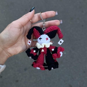 May include: A crochet jester keychain featuring a white face with black and red eye markings, a cream-colored ruffled collar, and a body in alternating black and red sections. Small playing card charms hang from the jester's hat.