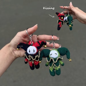 Puede incluir: Dos llaveros de bufón de ganchillo, uno en rojo y negro y otro en verde oscuro, cada uno con cara blanca, gorro puntiagudo con cascabeles dorados y cuello con volantes. Un recuadro muestra a los dos bufones juntos con el texto Kissing.