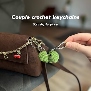 May include: Two small green crochet frog keychains attached to a brown suede bag with a gold chain featuring a red cherry charm. The text Couple crochet keychains Ready to ship is visible above the bag. The frogs are positioned facing each other.