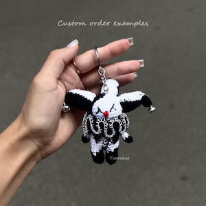 Puede incluir: Un llavero de bufón tejido a ganchillo en blanco y negro con una pequeña nariz roja, ojos en forma de X y un cuello con volantes. La figura tiene cascabeles plateados en las puntas del gorro y un anillo metálico. Texto: Custom order examples.