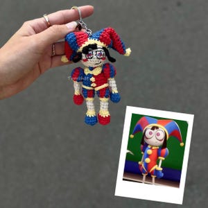 Puede incluir: Un llavero de ganchillo con forma de bufón que luce un gorro rojo, azul y amarillo, un conjunto a juego con mangas abullonadas y pelo oscuro. La figura tiene rasgos faciales bordados y está unida a una anilla de metal plateado. Texto Tvorchet visible.