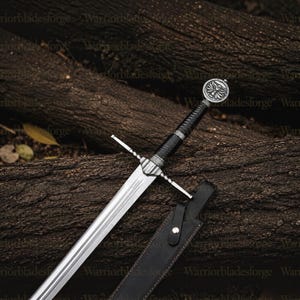 May include: A silver-toned steel sword featuring a black wrapped grip, a crossguard with twisted metal detailing, and a circular pommel embossed with a winged emblem. The sword is accompanied by a black leather scabbard with a metal snap closure.