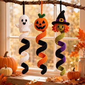 May include: Three hanging crochet ornaments featuring a white ghost, an orange jack-o'-lantern, and a witch with a black hat and orange hair. Each figure has a long, spiralling tail in alternating colours, suspended from a branch by black or orange loops.