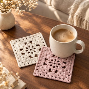 May include: Two square crochet coasters featuring a central floral motif with intricate openwork patterns. One coaster is off-white and the other is dusty pink. A ceramic mug filled with coffee sits on the pink coaster, showcasing the texture.