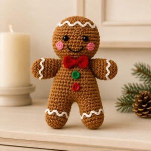 Puede incluir: Un muñeco de ganchillo marrón con forma de hombre de jengibre que presenta una pajarita roja, un botón verde, un botón rojo, mejillas rosas, ojos negros y detalles de glaseado blanco ondulado en la cabeza, las muñecas y los tobillos.