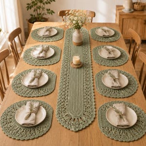 Può includere: Un runner da tavola all'uncinetto color verde salvia e sei tovagliette rotonde abbinate su un tavolo in legno. Ogni tovaglietta presenta un piatto in ceramica bianca sormontato da un tovagliolo di lino beige piegato e un rametto verde.