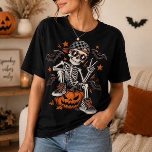 Op de afbeelding: Een zwart t-shirt met een afbeelding van een skelet met een geblokte baseballpet en een oranje zonnebril, zittend op een pompoen. Het skelet maakt een vredesteken en wordt omringd door oranje sterren en zwarte vleermuizen op de voorkant.