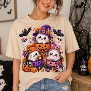 Peut inclure: Un t-shirt beige à manches courtes avec une impression graphique colorée représentant trois fantômes de dessin animé en costumes d'Halloween. Les fantômes sont assis dans des seaux citrouilles orange et violets entourés de chauves-souris.