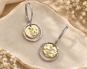 May include: A pair of drop earrings featuring a silver-toned hoop and a smaller, circular gold-toned charm with a hammered texture and an engraved leaf design. The earrings are attached to silver-toned lever-back closures for secure wear.