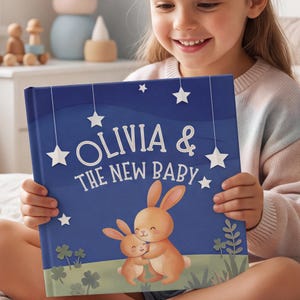 Può includere: Un libro per bambini con copertina rigida blu scuro che riporta il titolo Olivia & The New Baby in bianco. L'illustrazione mostra due coniglietti marroni che si abbracciano in un prato sotto stelle bianche sospese nel cielo notturno.