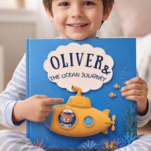 Könnte beinhalten: Ein gebundenes Buch mit dem Titel Oliver & The Ocean Journey mit blauem Einband und einem gelben U-Boot. Ein gezeichneter Bär mit Matrosenmütze ist durch das Fenster des U-Boots zu sehen. Das Cover zeigt zudem kleine Fische und Seetang.