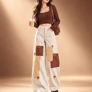 Puede incluir: Un top corto marrón de manga larga con escote cuadrado y botones, combinado con jeans de pierna ancha y talle alto en color crema con parches de tela marrón y beige y detalles de flores bordadas, usado con zapatos de plataforma marrones.