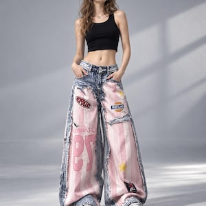 Può includere: Un top corto nero senza maniche abbinato a jeans a gamba larga che presentano un mix di denim blu lavaggio acido e pannelli a righe verticali rosa chiaro. I jeans includono bordi sfrangiati, varie toppe colorate e il numero 97 stampato in rosa.