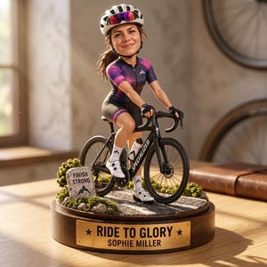 Könnte beinhalten: Eine individuelle Wackelkopf-Figur eines Radfahrers mit weißem Helm, lila-schwarzem Trikot und schwarzer Hose auf einem schwarzen Rennrad. Der Sockel zeigt einen Stein mit der Aufschrift FINISH STRONG und ein Schild mit RIDE TO GLORY SOPHIE MILLER.