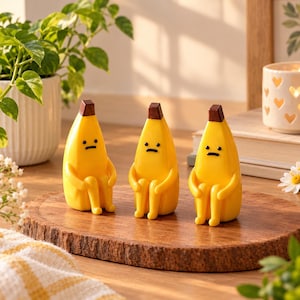 May include: Three yellow banana figurines with brown stems, each featuring a simple black line for a mouth and two black dots for eyes. The figures are depicted in a seated position with arms resting on their knees, showcasing a smooth, glossy finish.