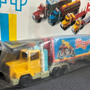 Könnte beinhalten: Ein gelb-blauer Spielzeug-Sattelschlepper mit einem Anhänger, der eine Grafik einer Person auf einem Motorrad und den Text Hot Cycle zeigt. Der LKW ist in einer durchsichtigen Plastiktüte versiegelt und auf einer Papprückwand befestigt.