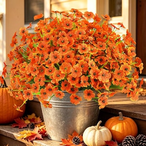 May include: A large, dense arrangement of vibrant orange artificial flowers with green stems, housed in a rustic, cylindrical galvanized metal bucket. The flowers feature five-petaled blooms with textured surfaces and dark centers.
