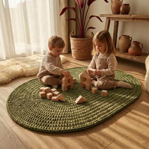 Könnte beinhalten: Ein ovaler, olivgrüner Häkelteppich aus dickem Garn. Zwei Kinder sitzen auf dem Teppich und spielen mit einem Set aus hellen Holzbausteinen. Der Teppich hat eine strukturierte, geschlungene Oberfläche und liegt auf einem Holzboden.