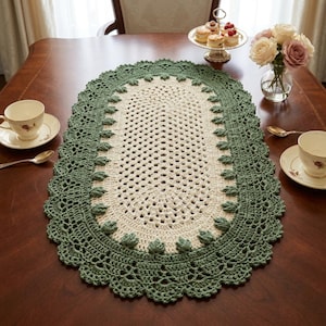 Peut inclure: Un chemin de table ovale en crochet avec un centre couleur crème au motif ajouré répétitif, entouré d'une large bordure festonnée vert sauge agrémentée de petits motifs en relief en forme de feuilles sur le bord intérieur.
