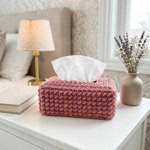 May include: A rectangular tissue box cover crafted from dusty rose yarn with a textured, crocheted stitch pattern. The cover features an open top design that allows white facial tissues to be pulled through the center opening with ease.