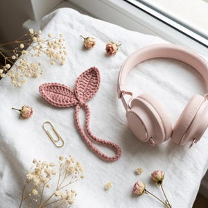 May include: A pair of light pink over-ear headphones, a dusty rose crocheted leaf bookmark with a long stem, a gold metal paperclip, and several small dried flowers arranged on a white fabric surface with a visible woven texture.
