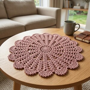 Può includere: Un centrino all'uncinetto circolare color rosa polvere, caratterizzato da un intricato motivo radiale traforato con bordo smerlato. Il pezzo tessile è realizzato con filato sottile e presenta un design simmetrico e dettagliato.