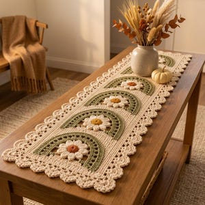 Peut inclure: Un chemin de table rectangulaire en crochet couleur crème, orné d'un motif répétitif d'arches en demi-cercle vert sauge et de fleurs de marguerite blanches à cœur jaune. Le chemin présente un bord festonné et un aspect texturé fait main.