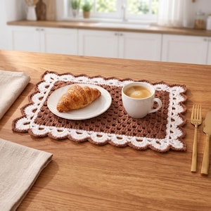 May include: A rectangular brown crochet placemat with a white scalloped border. A white ceramic plate holds a golden-brown croissant, and a white ceramic mug filled with coffee sits on the placemat next to a gold fork and knife.